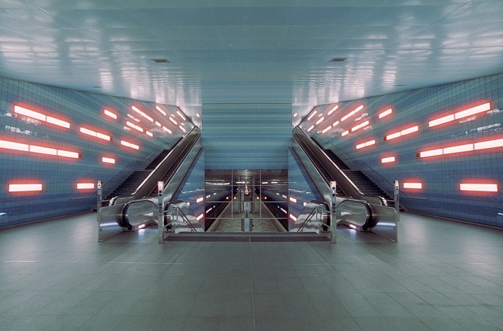 Two escalators lead down to a subway platform with blue tiled walls and orange lights.
