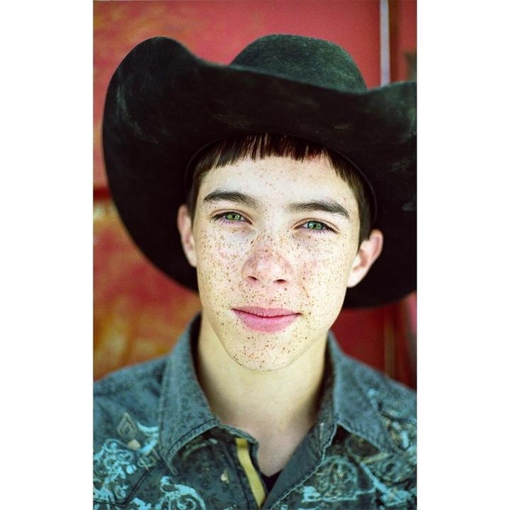 A boy with freckles and green eyes wears a cowboy hat and a patterned shirt.
