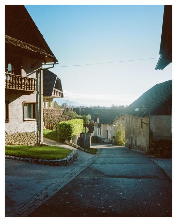 A narrow street winds through a village with houses and green hedges under a clear sky.