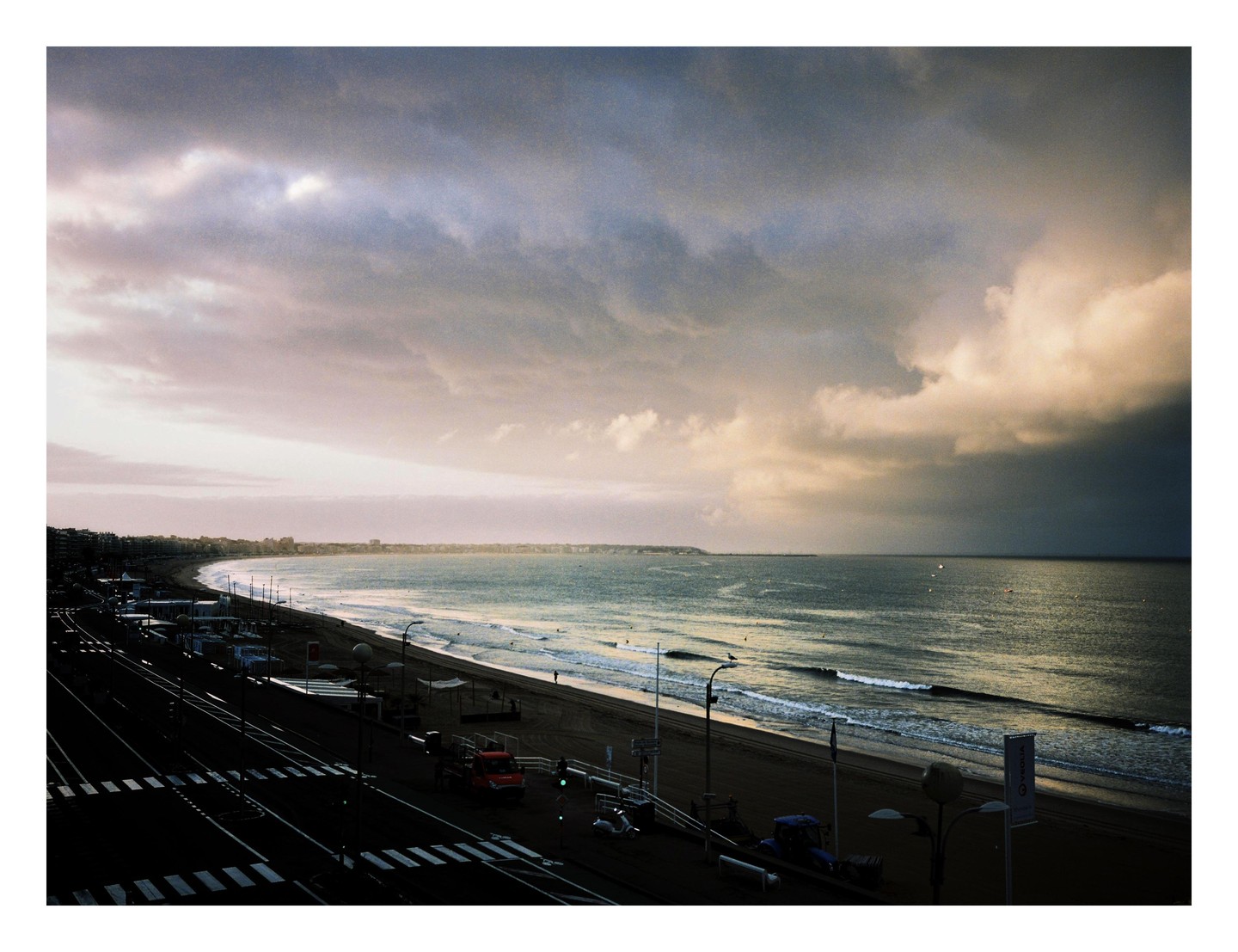A wide beach with a road and buildings runs along a bay under a dramatic, cloudy sky.