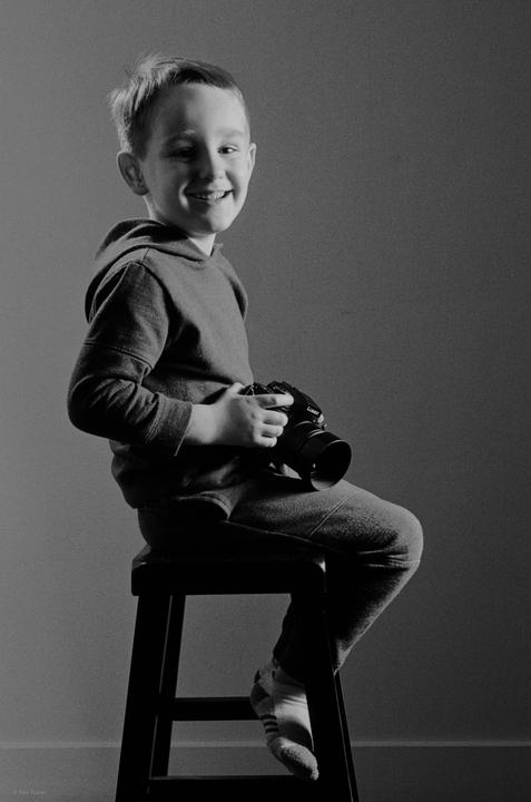 A smiling boy sits on a stool holding a camera.