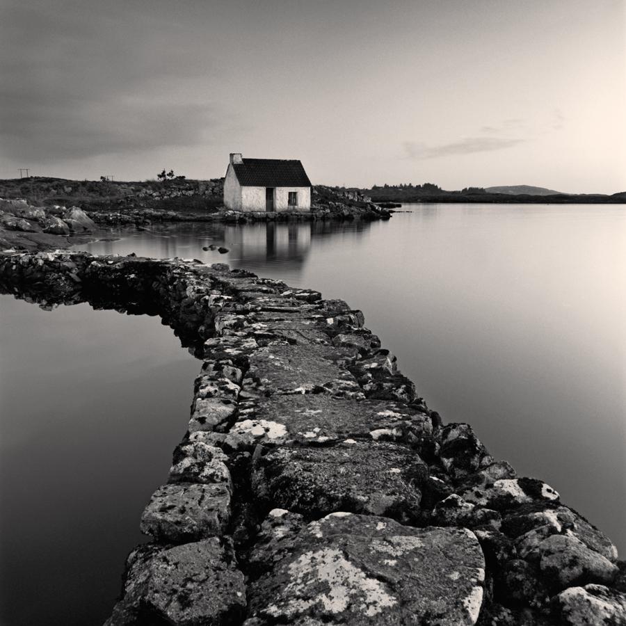 A stone path leads to a small house on the shore of a calm lake under a cloudy sky.