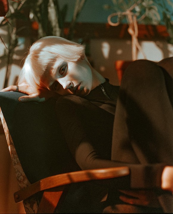 A woman with short blonde hair reclines in a chair, looking towards the viewer.