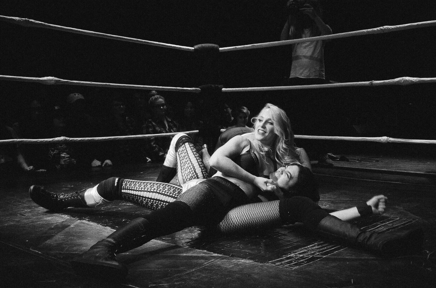 Two women wrestle in a ring as spectators watch in the background.