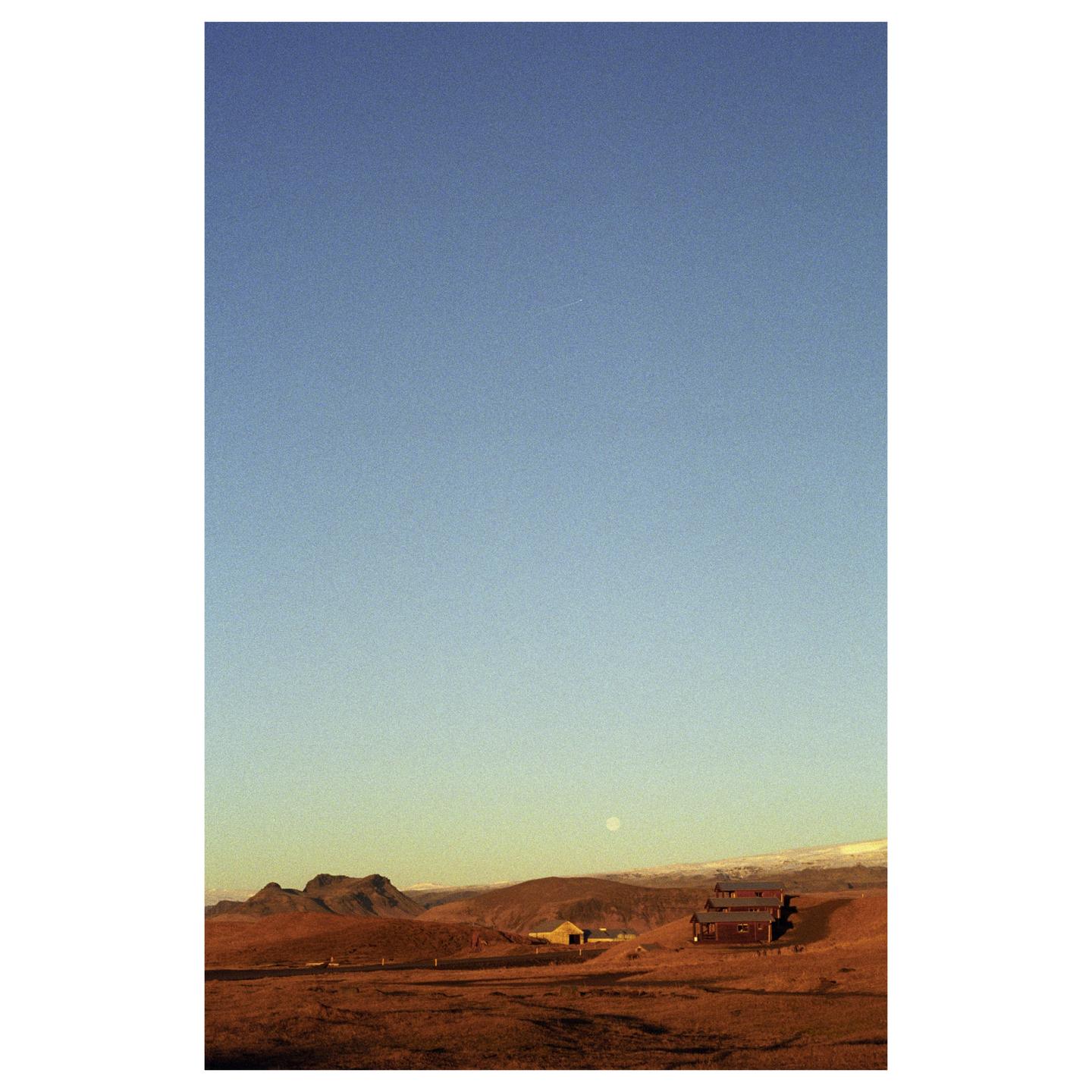 A red house and a small building sit in a vast, arid landscape under a clear sky with a visible moon.