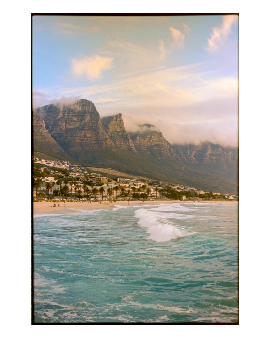A wave breaks on the shore of Camps Bay with mountains and a town in the background at sunset.