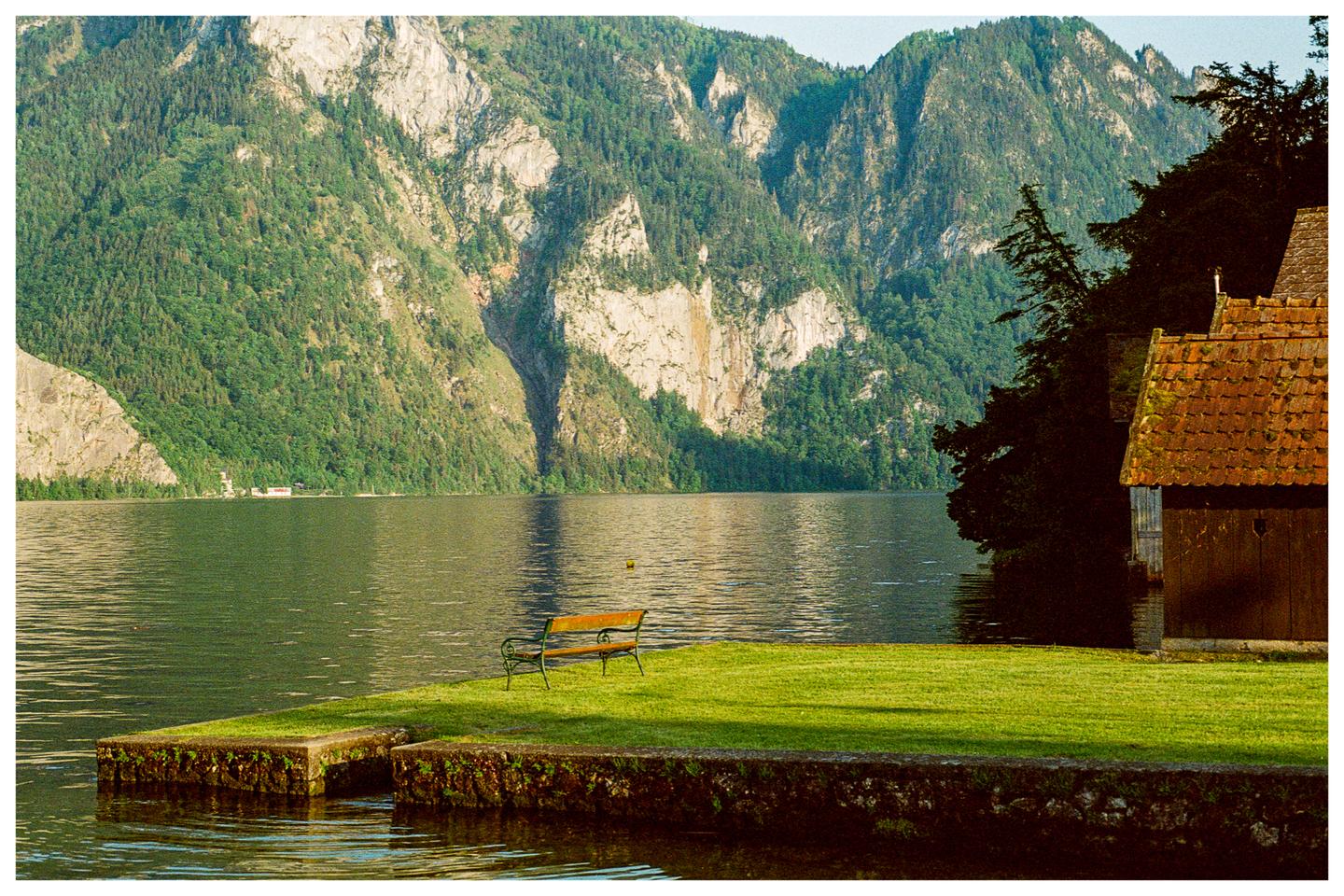 A wooden bench sits on a grassy bank next to a lake with mountains in the background.