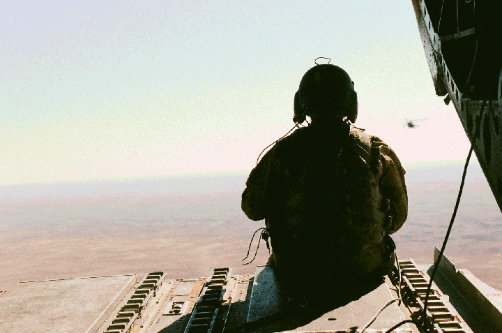 A person in military gear sits at the open rear of an aircraft, looking out over a vast landscape with another aircraft in the distance.