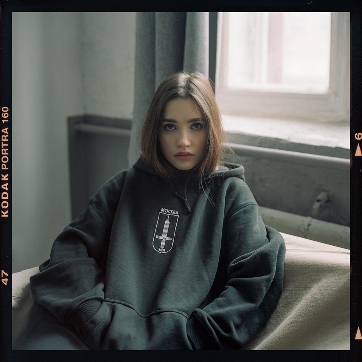 A woman wearing a dark hoodie with a white emblem sits indoors, looking directly at the viewer.