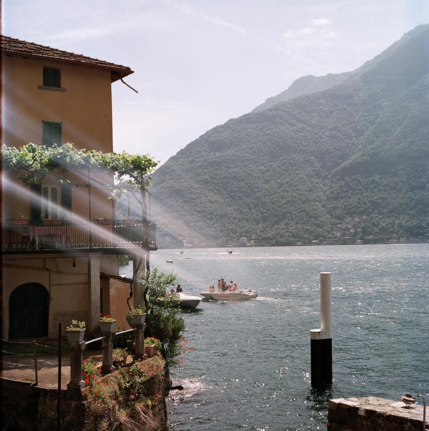 Sunlight streams over a building with a balcony overlooking a lake with boats and a mountain in the background.
