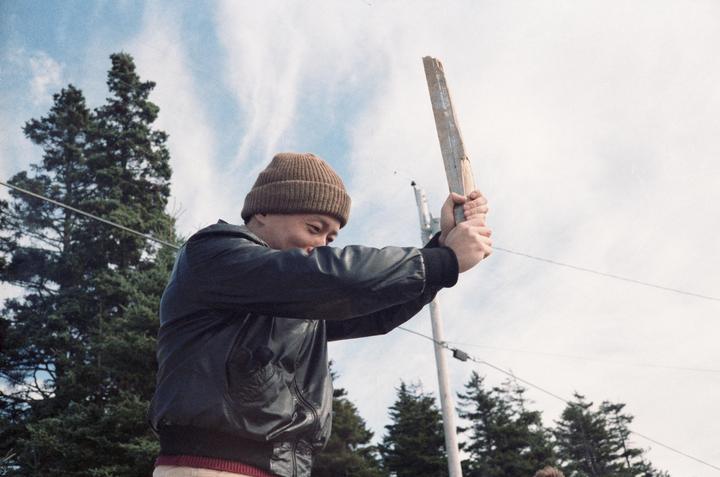 A man in a beanie and jacket holds a wooden plank against a cloudy sky with trees and utility poles in the background.