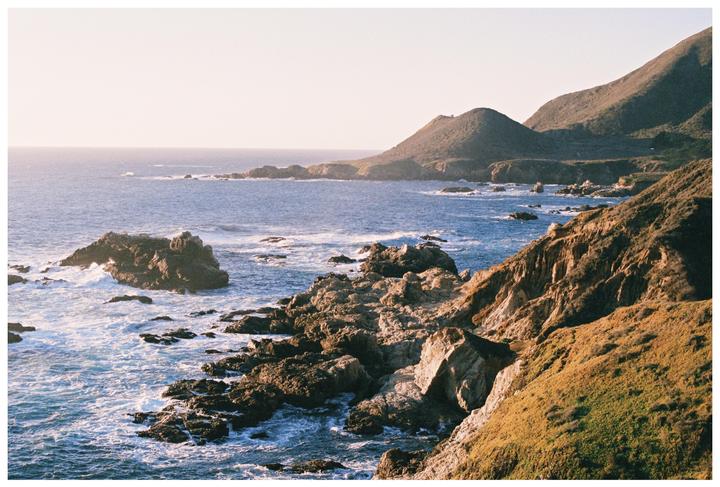 Waves crash against a rocky coastline with mountains in the background under a bright sky.