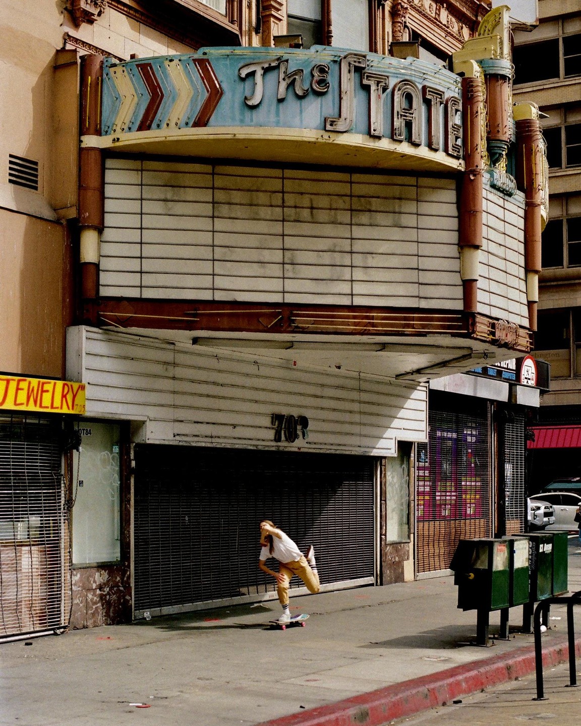 A person on a skateboard rides past a closed theater with a marquee that reads "The State" and several storefronts with metal security gates.