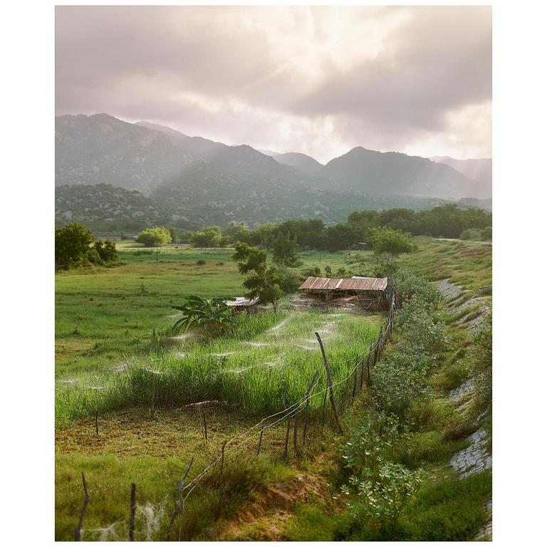 A small shack with a rusty roof sits in a green field with mountains in the background under a cloudy sky.