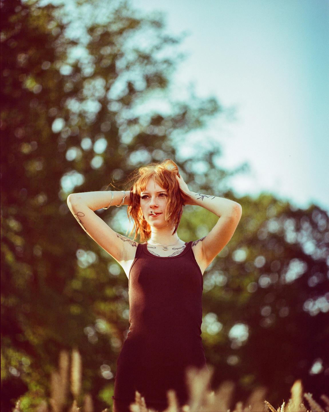 A woman with red hair and tattoos stands in a field with her hands behind her head.