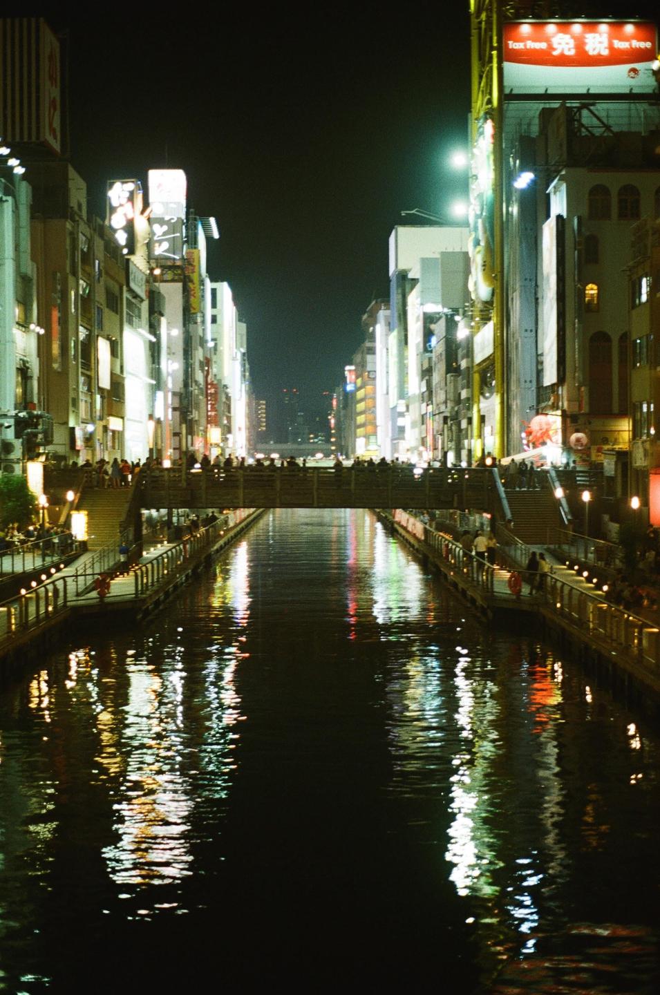 A canal with a bridge full of people is surrounded by buildings with bright signs at night.