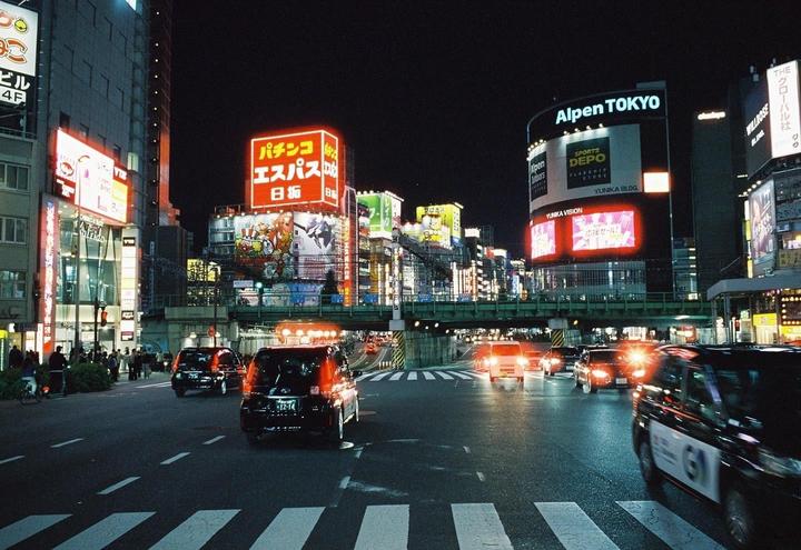 A busy street at night is filled with cars and illuminated by numerous neon signs and billboards.