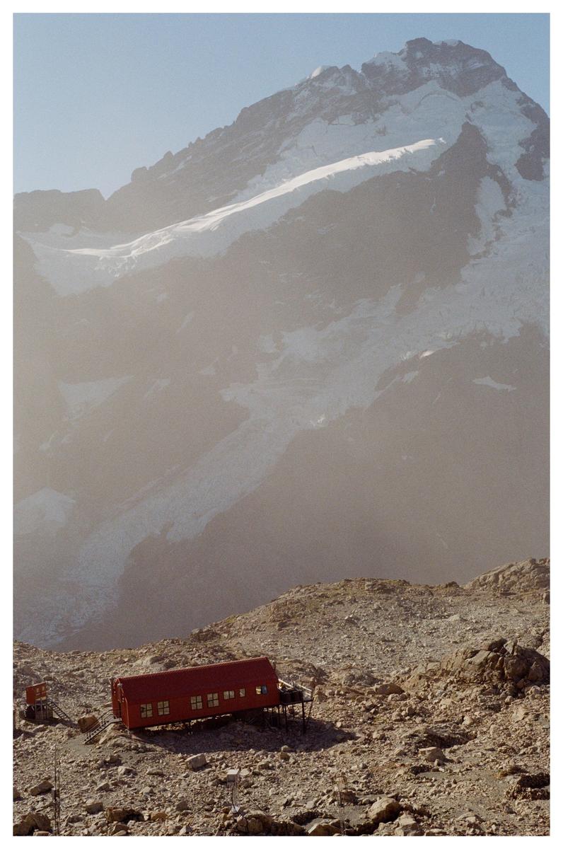 A red hut sits on a rocky mountain slope with a snow-capped mountain in the background.