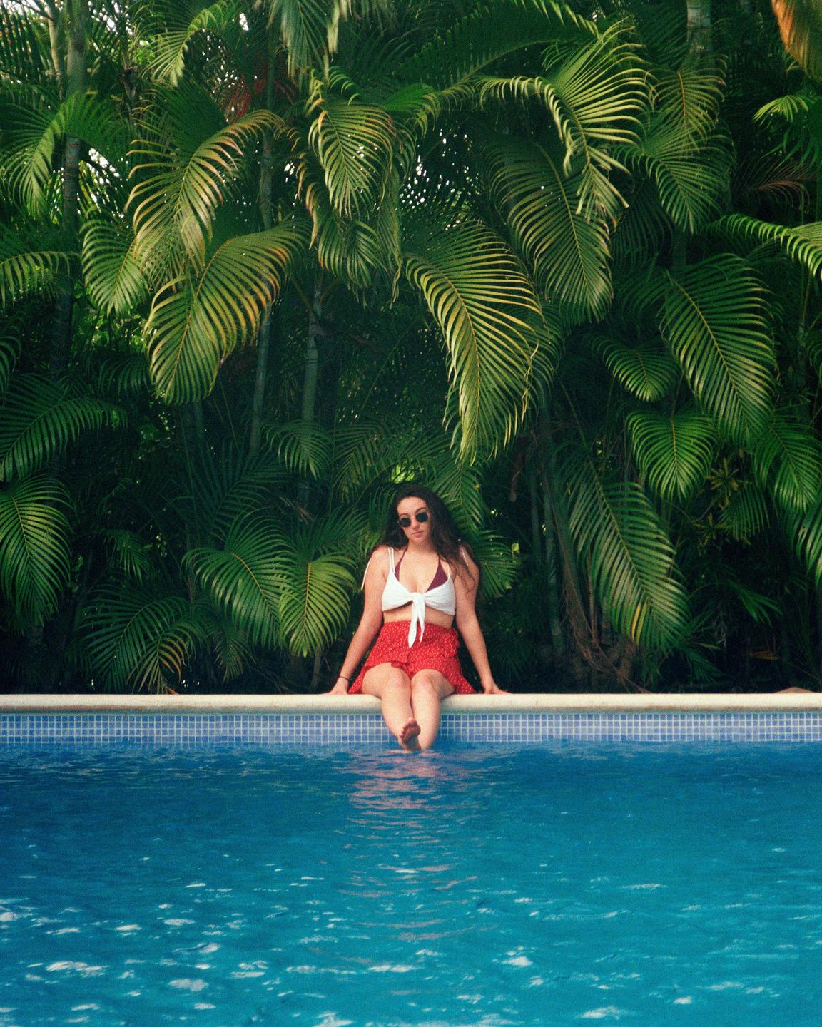A woman sits on the edge of a swimming pool with her feet in the water, in front of a dense background of palm fronds.