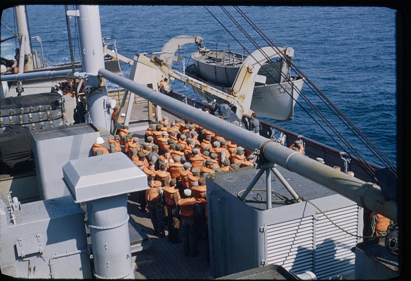 A group of men wearing life vests are gathered on the deck of a ship with a lifeboat visible in the background.