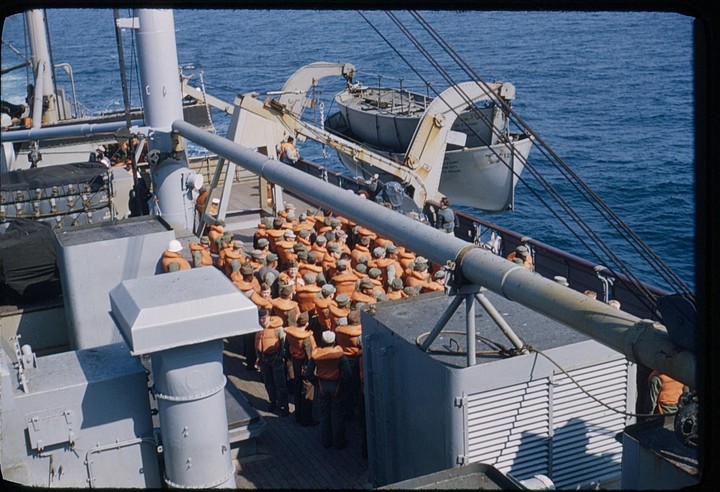 A group of men wearing life vests are gathered on the deck of a ship with a lifeboat visible in the background.