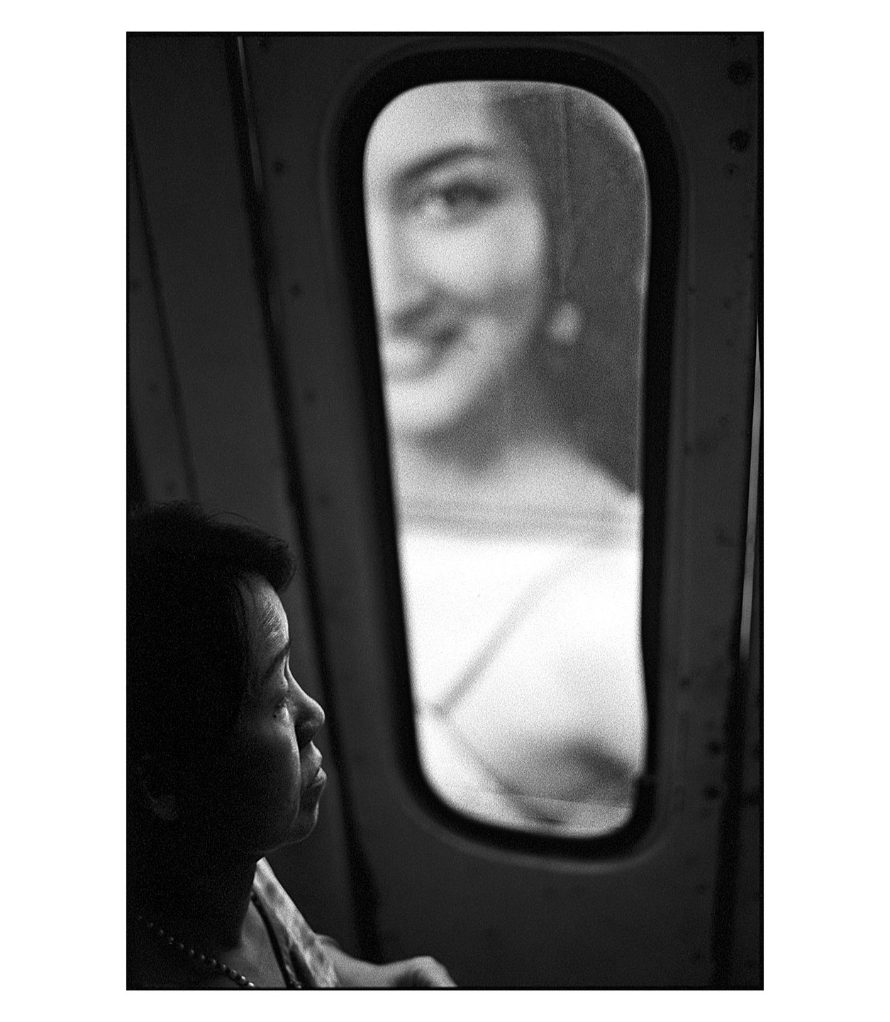 A woman looks out a bus window, with a reflection of a smiling woman visible in the glass.