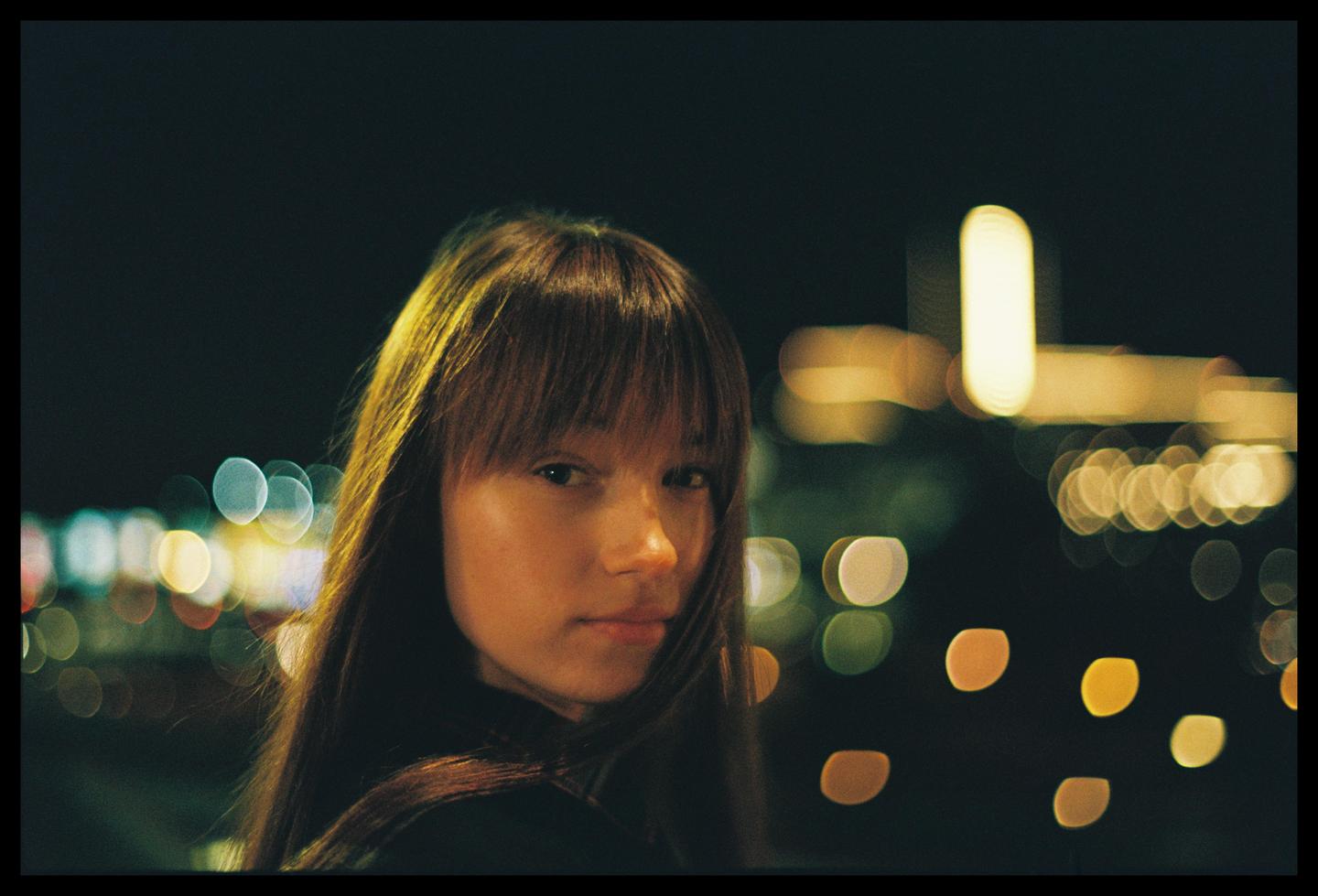 A woman with long hair and bangs looks at the viewer with blurred lights in the background.