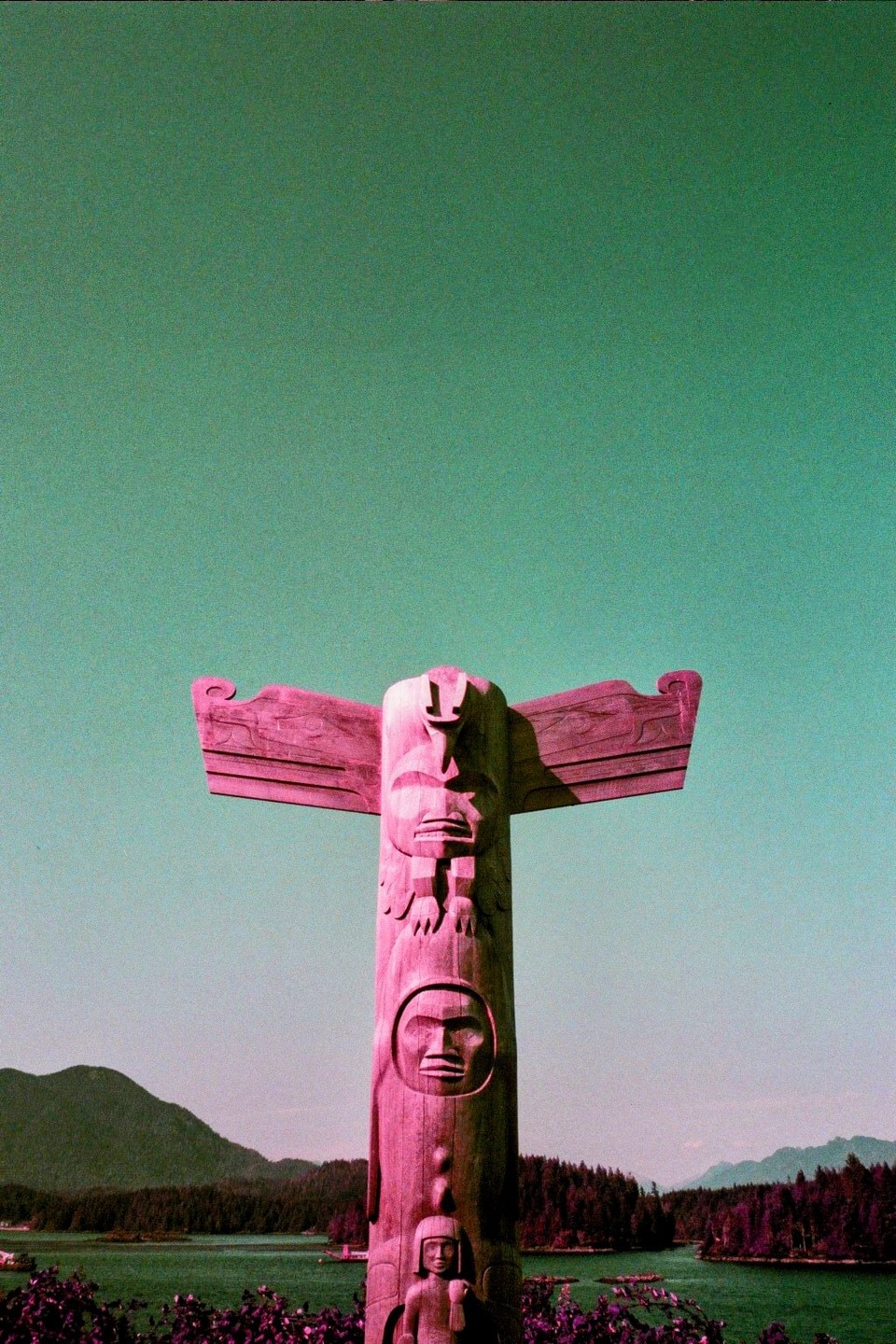 A totem pole stands tall against a green sky with mountains and water in the background.