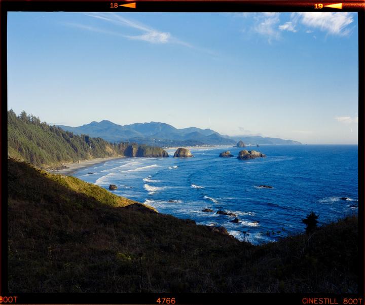 A coastal landscape features a beach, ocean, and mountains under a clear sky.