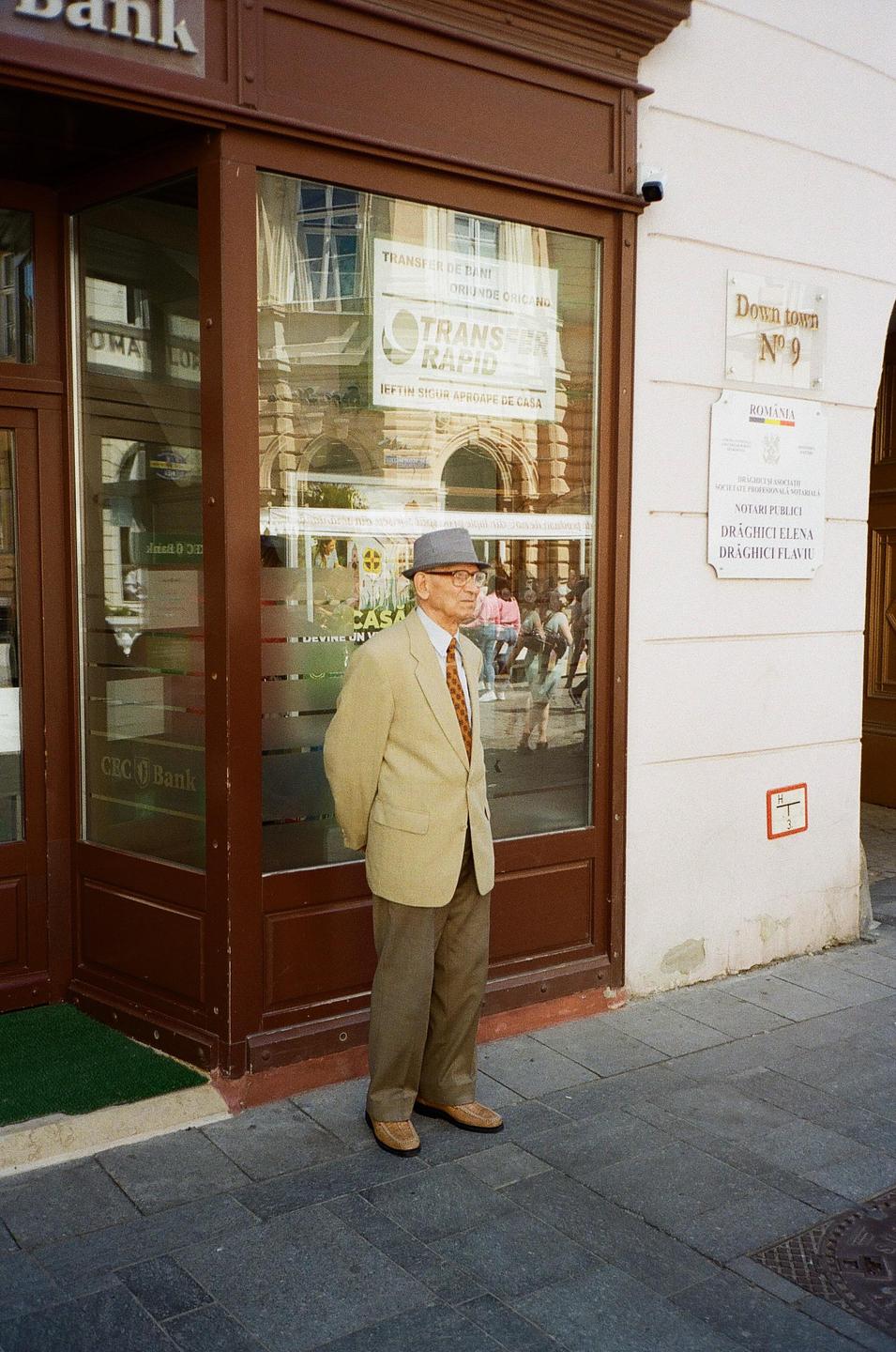 A man in a suit and hat stands on a sidewalk in front of a bank.