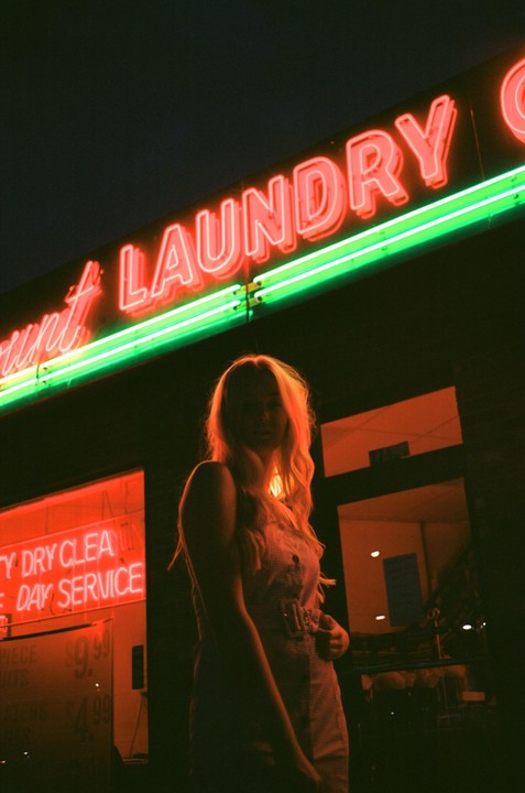 A woman stands in front of a laundry business with neon signs at night.