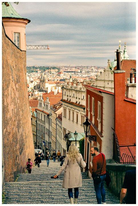 A woman and a man stand on a cobblestone street looking down at the city of Prague.