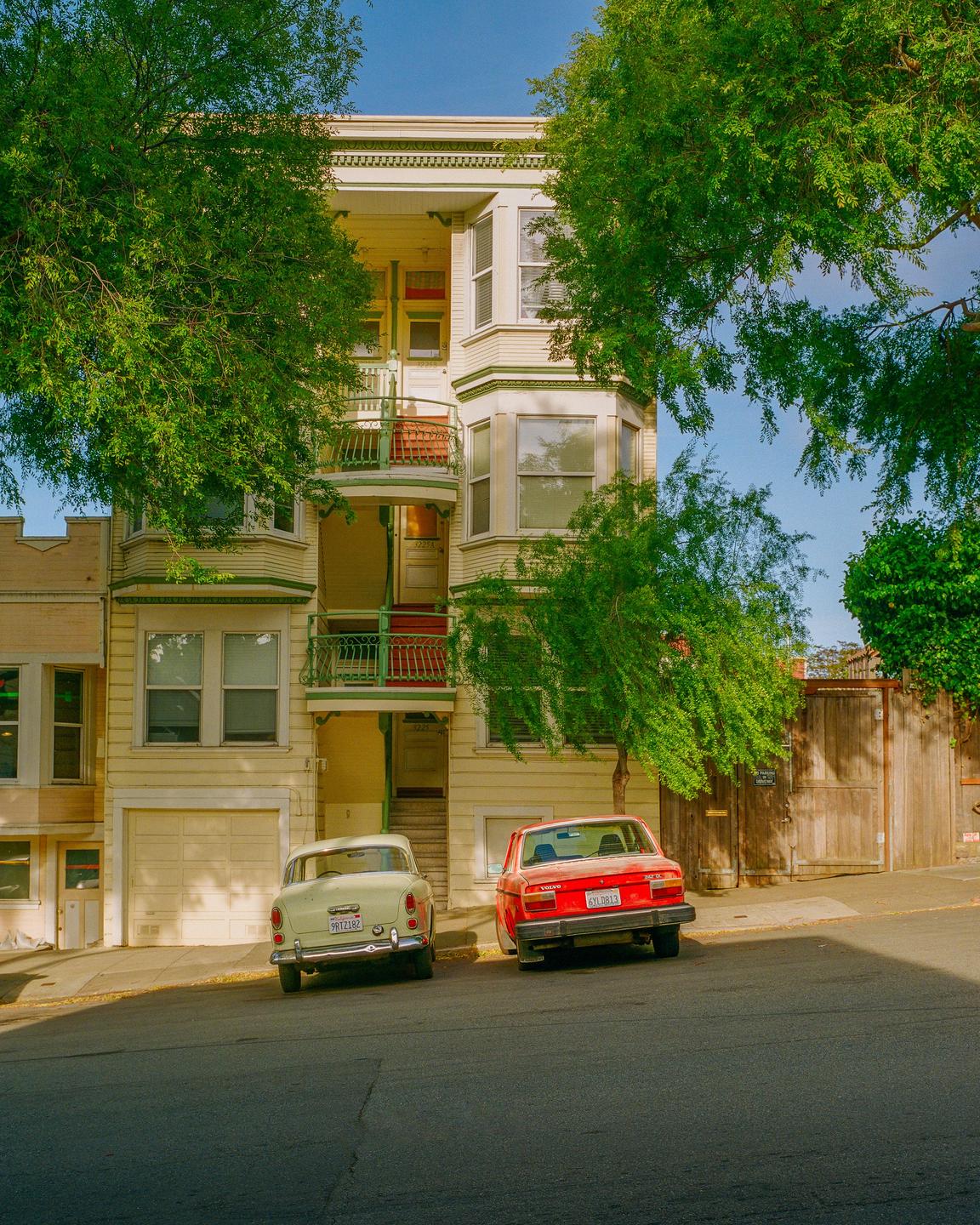 Two cars are parked on a sloped street in front of a multi-story building with bay windows and balconies.