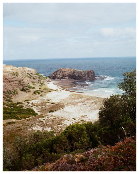 A coastal landscape features a sandy beach, rocky outcrops, and the ocean under a cloudy sky.