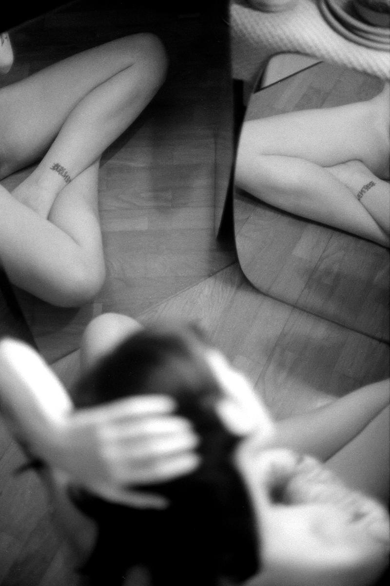 A woman's legs with a tattoo on her ankle are reflected in a mirror, with her head and arms out of focus in the foreground.