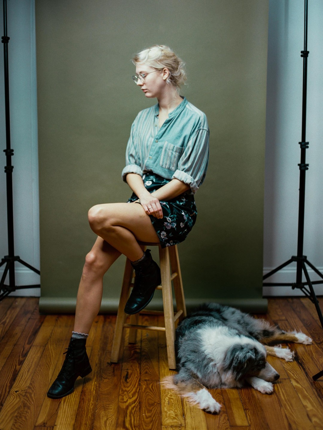 A woman sits on a stool with a dog lying on the wooden floor next to her.