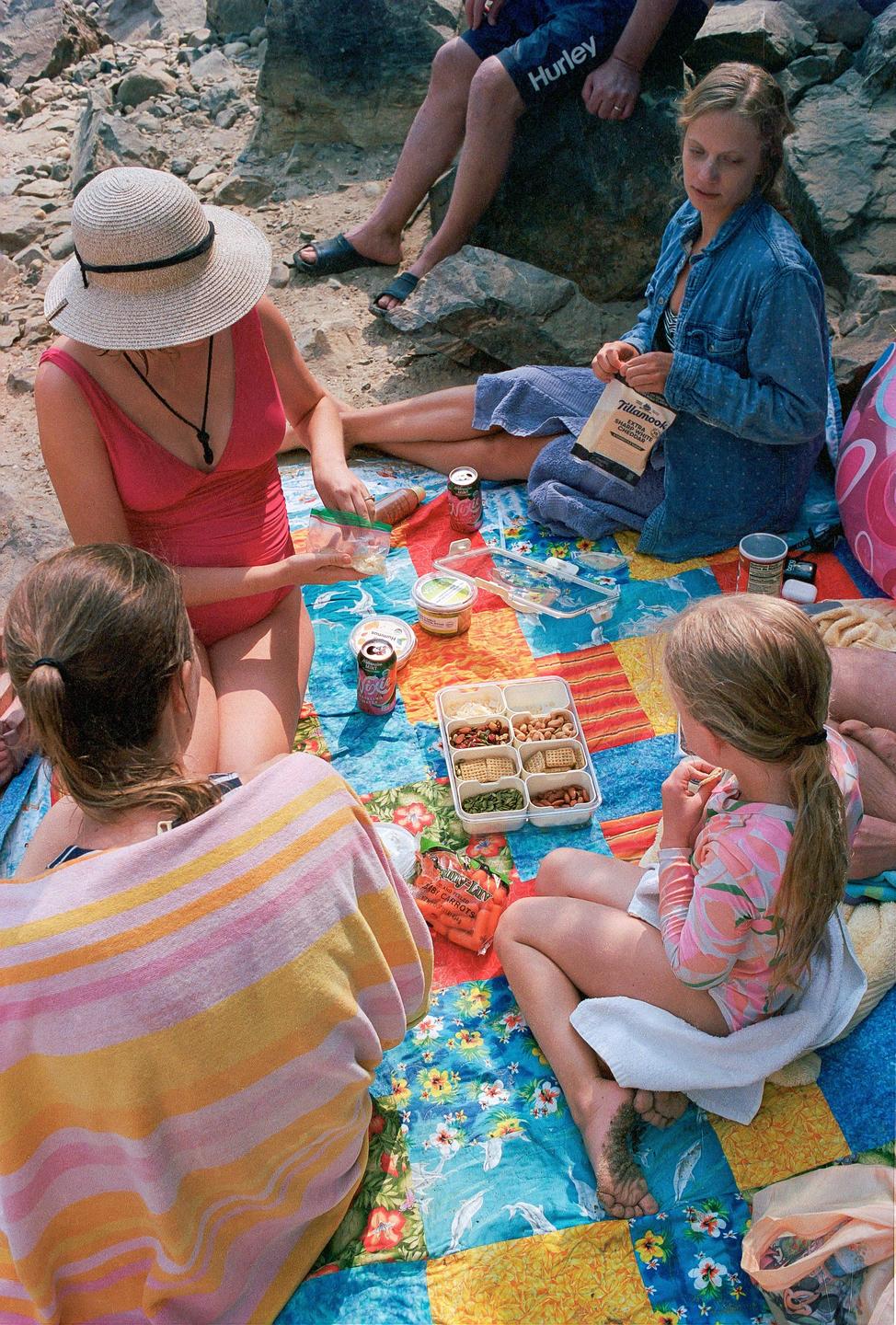 A family is having a picnic on a colorful blanket on a rocky beach.