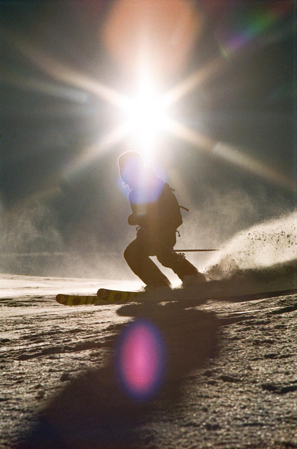 A skier is captured in silhouette against a bright sunburst, kicking up snow as they move down a slope.