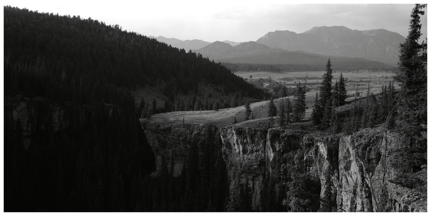 A panoramic landscape shows a valley with mountains in the distance, framed by a dark, forested cliff on the left and a sunlit cliff on the right.