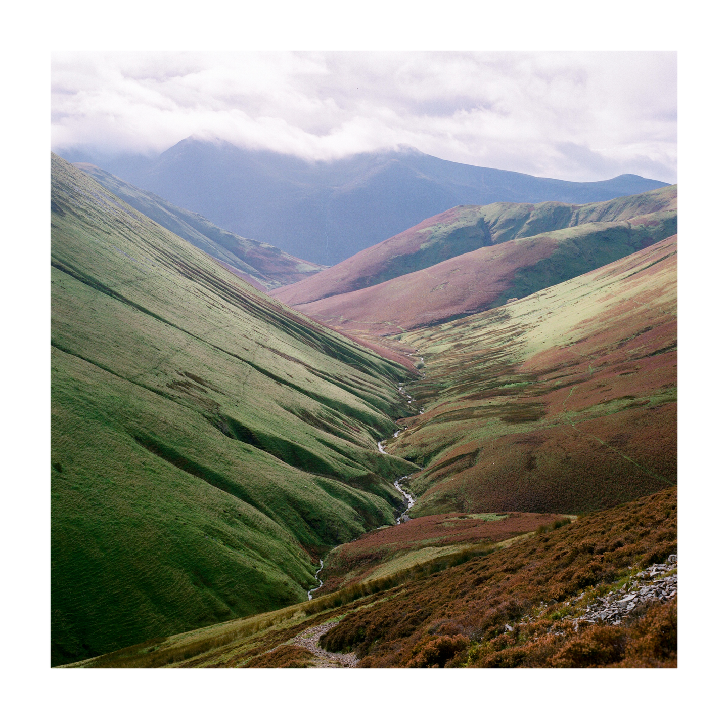 A narrow stream winds through a valley with steep, grassy slopes under a cloudy sky.