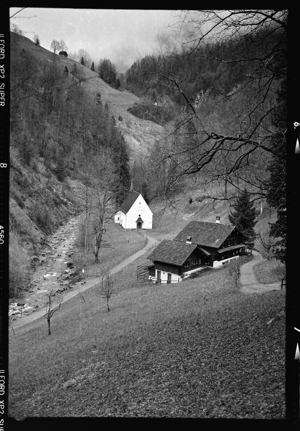 A small white church and a house with a tiled roof are nestled in a valley next to a river.
