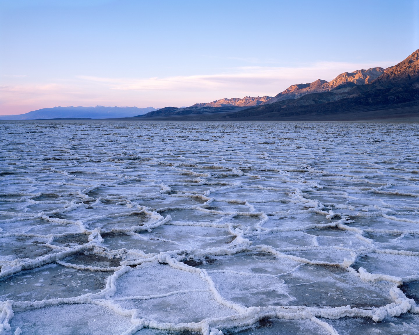 A vast salt flat with polygonal patterns stretches towards distant mountains under a pastel sky.