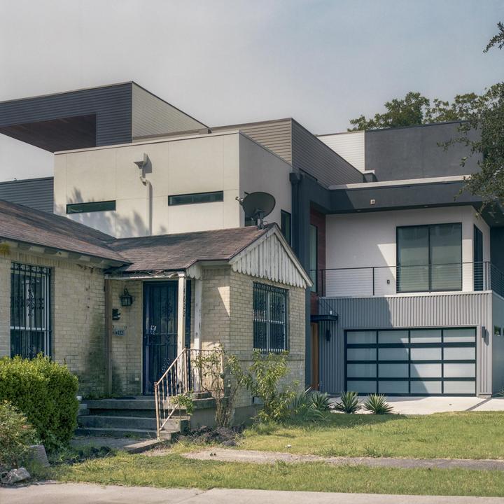 An old brick house with a porch and security bars stands next to a modern house with a garage and balcony.
