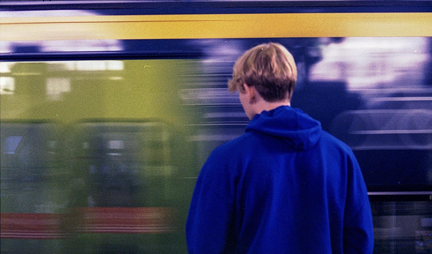 A person in a blue hoodie stands with their back to the camera as a train passes by.