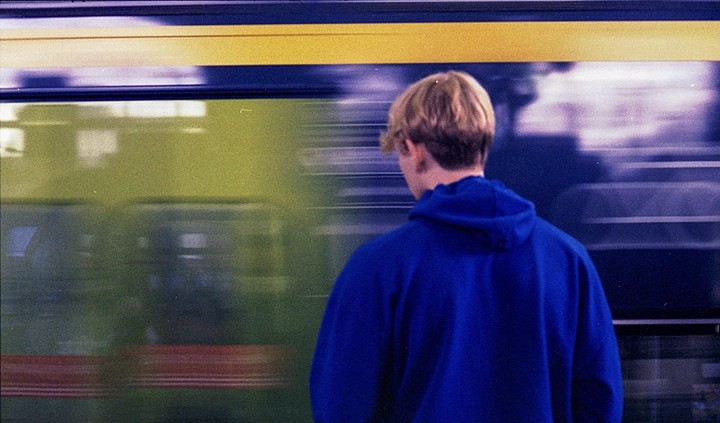 A person in a blue hoodie stands with their back to the camera as a train passes by.
