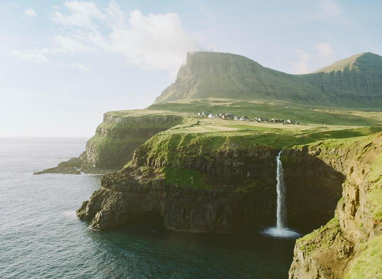A waterfall cascades from a green cliff into the ocean, with a village nestled on a distant plateau under a large mountain.