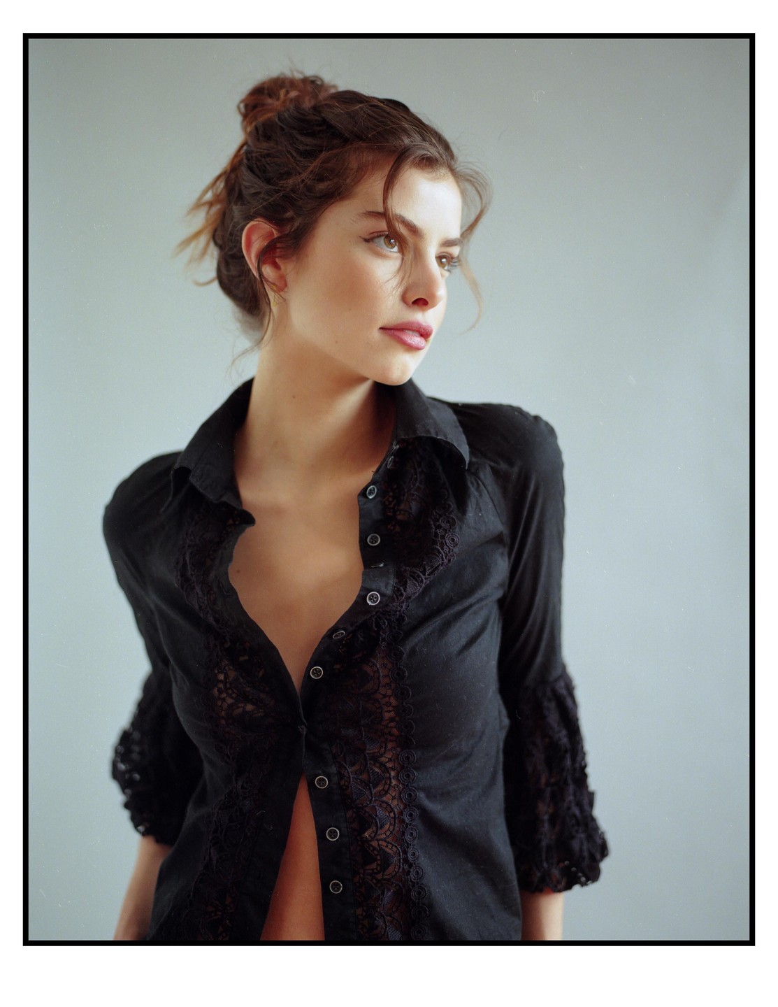 A woman with her hair in a bun wears an unbuttoned black lace shirt and looks to the right.