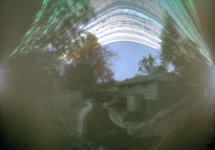 A long exposure image shows a house and trees under a bright sky with light trails.