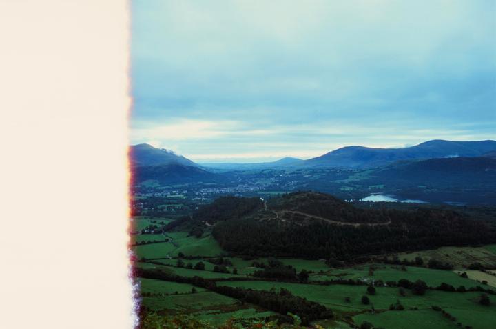 A wide landscape shows a valley with a town, a lake, and mountains under a cloudy sky, with a large light leak on the left side.