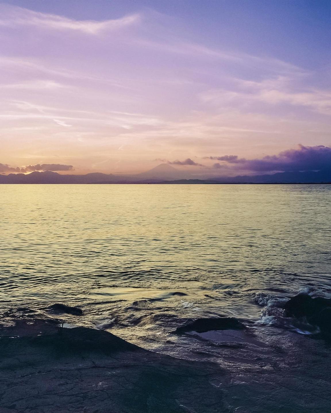 A body of water with rocks in the foreground reflects the colorful sky at sunset, with mountains visible in the distance.